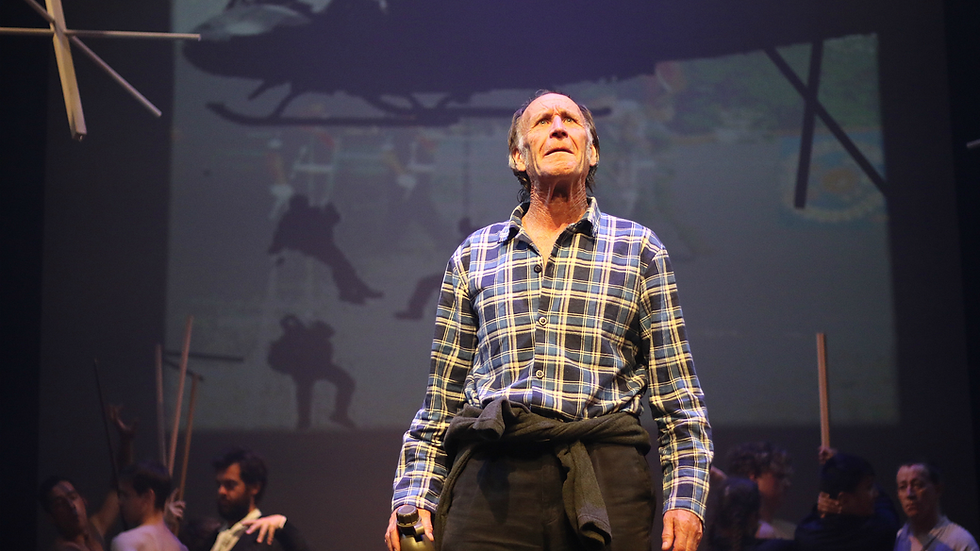 An older man stares up into the rafters, a war scene with a helicopter plays out behind him