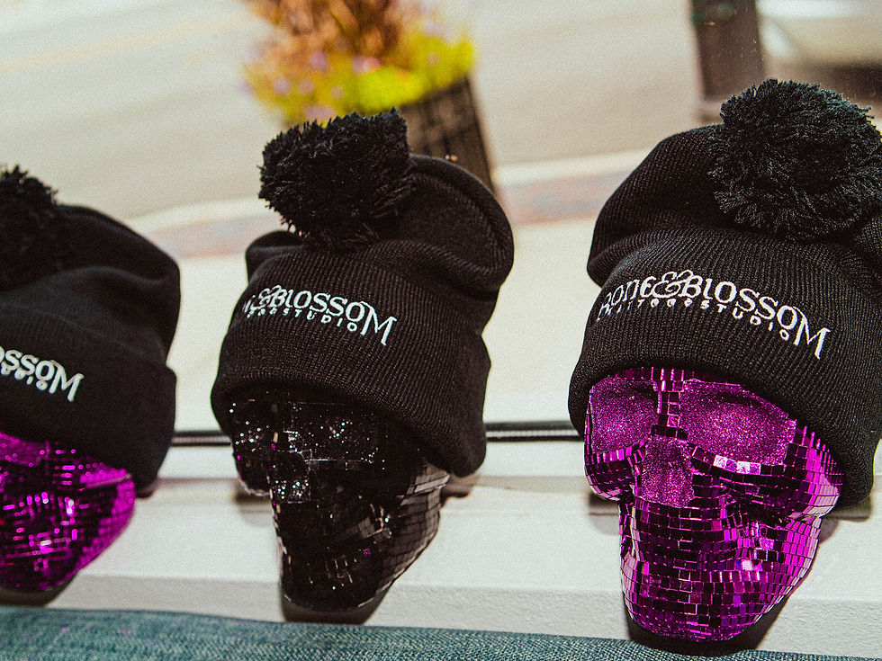 Bone & Blossom Tattoo Studio black knit beanies with pom-poms displayed on glittery purple and black skull decorations in a shop window.