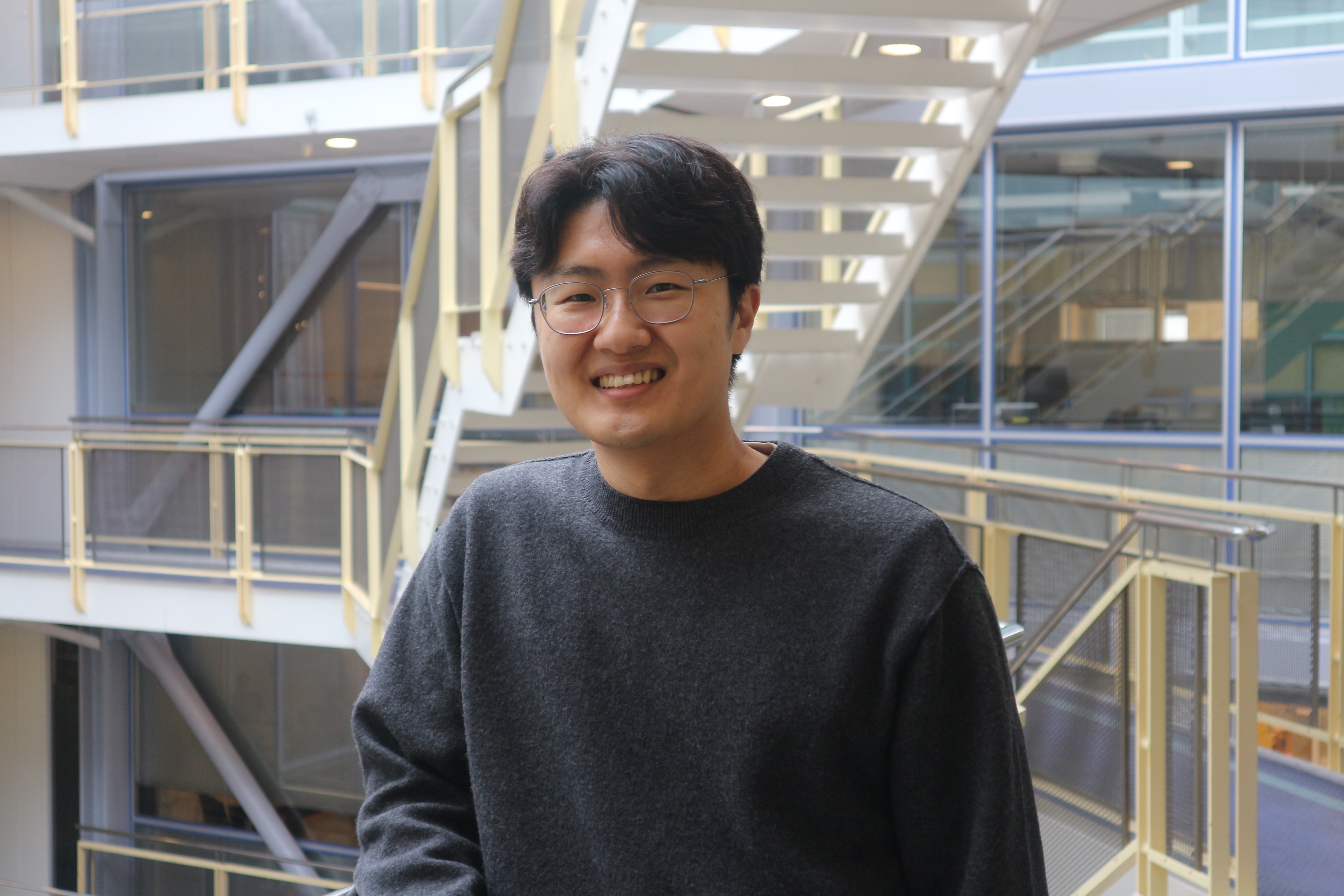 Picture of Dongkuyn Lim standing in front of an open staircase. He is smiling, has dark hair, wears glasses and a dark grey jumper.