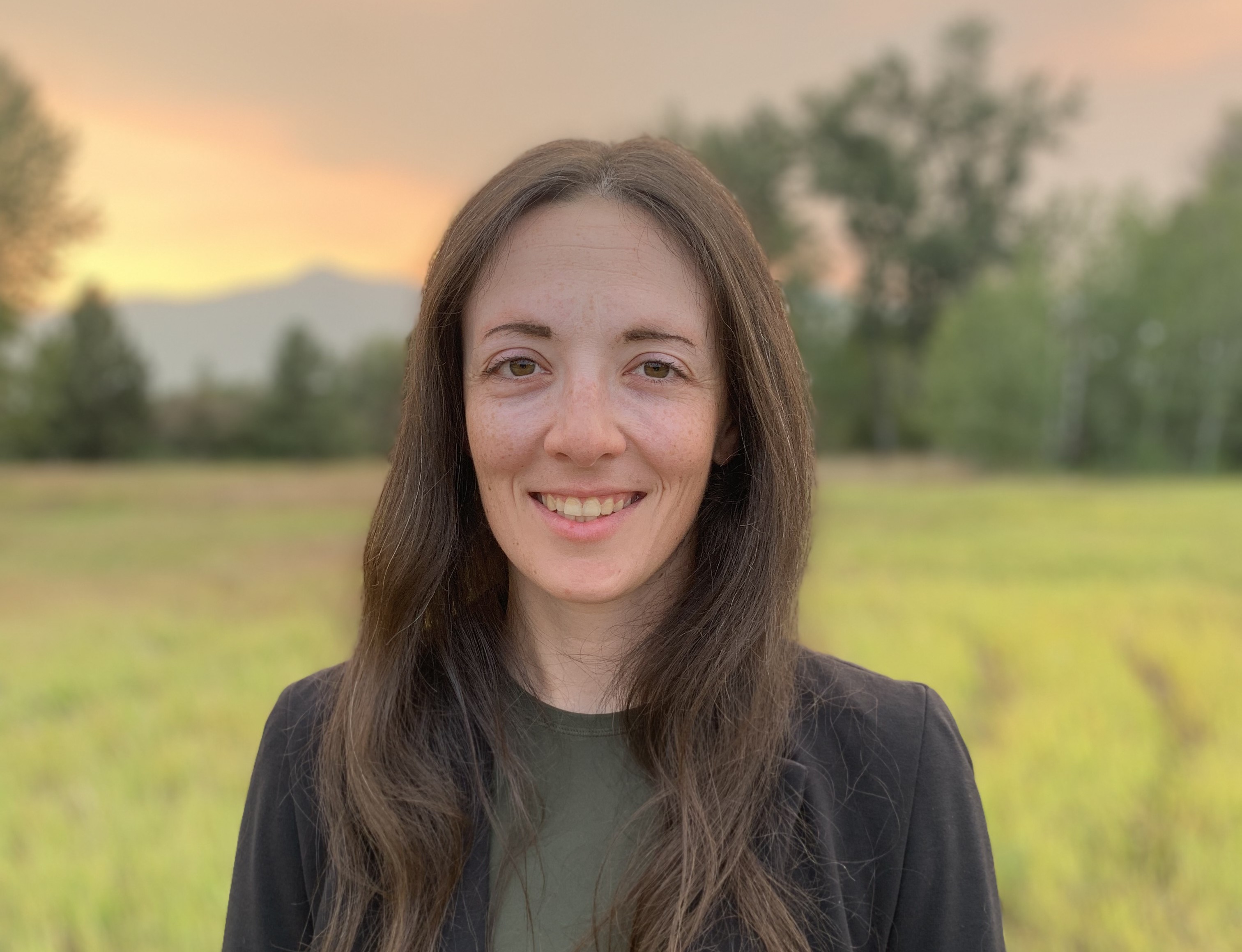 Portrait of Meaghan with long brown hair with grass, trees and the sun setting over the mountain in the background.