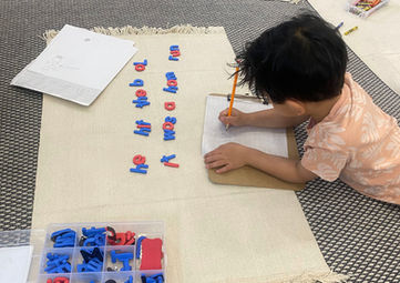 Child is laying down on the floor drawing the letters he sees in a Montessori classroom.