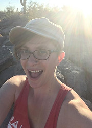 Sarah is a white, androgynous and queer woman with short hair and smiling at the camera. They are wearing a red tank top, glasses, and a brown cap. The background is the Sonoran Desert and the sun is shining from the top right corner of the picture creating rays of sunshine towards their face and the camera.