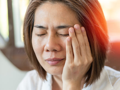 Woman with brown hair, eyes closed, touching her face with one hand. She appears in discomfort from jaw pain. Sunlight filters in the background.