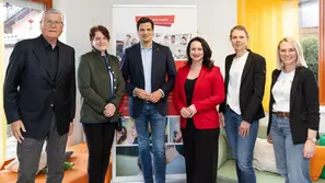 Sechs Personen posieren lächelnd vor einem Banner in einem Raum mit gelben Vorhängen. Eine Frau trägt ein rotes Jackett.