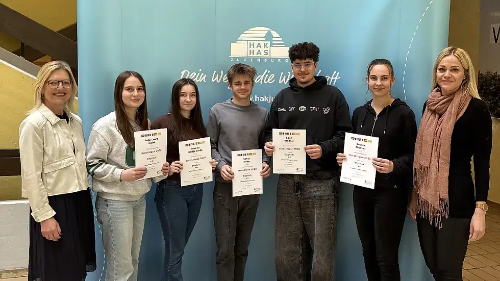 Sieben Personen halten Zertifikate vor einem türkisfarbenen Hintergrund mit "HAK HAS Judenburg" Logo. Sie wirken stolz und froh.