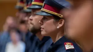 Profil einer uniformierten Frau mit Mütze und Schulterabzeichen bei einer Zeremonie, unscharfe Kollegen im Hintergrund.