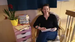 Birgit Krenn sitzt mit Notizblock und Stift in einem gemütlichen Raum neben einem Stapel Bücher und einer Pflanze und leitet Schreibwerkstätten im Rahmen von „Erlebnis Sprache“ in Fohnsdorf.