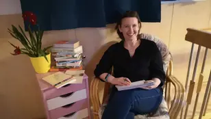Birgit Krenn sitzt mit Notizblock und Stift in einem gemütlichen Raum neben einem Stapel Bücher und einer Pflanze und leitet Schreibwerkstätten im Rahmen von „Erlebnis Sprache“ in Fohnsdorf.