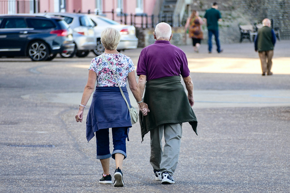 Elderly couple walks hand in hand down a paved street, backs to camera, with parked cars and other pedestrians in the background.