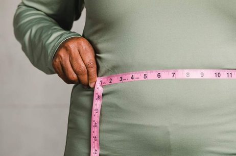 A person in a green shirt holds a pink measuring tape around their waist. The background is plain, highlighting the focus on measuring size.