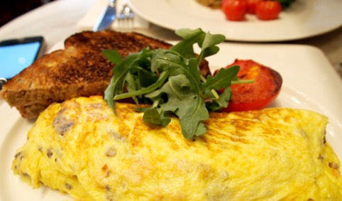 Omelette with rocket, toast, and grilled tomato on a white plate. Background shows phone and fork. Bright and appetizing setting.