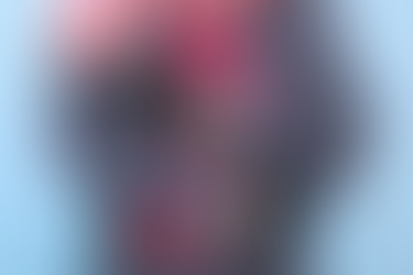 Person in gray sweater and gloves with floral design holds a red hot water bottle. Wearing a colourful striped scarf against a blue background.