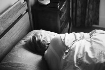 Black-and-white photo of a person sleeping on their side in bed, head on pillow, with a blanket in a quiet bedroom.