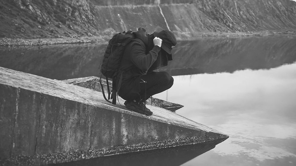 Person in black squats by water, looking down, on a concrete structure.