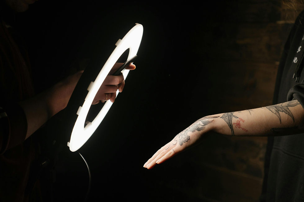 A close-up shot shows a person holding a large ring light, illuminating a tattooed arm reaching out from the right side of th