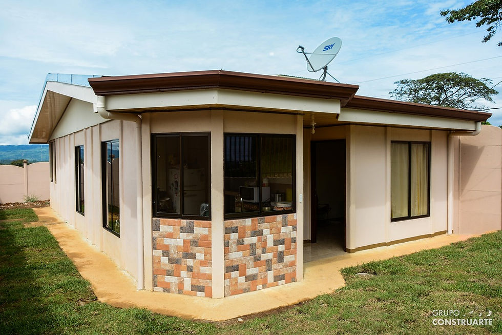 Casa beige con techo marrón, paredes decoradas con mosaico de ladrillos. Antena parabólica en el techo. Césped verde alrededor.