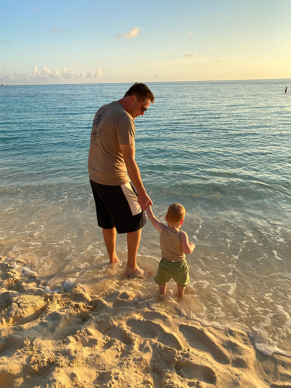 Adult and child holding hands, standing at the water's edge on a sandy beach during sunset. Calm ocean and clear sky in the background.