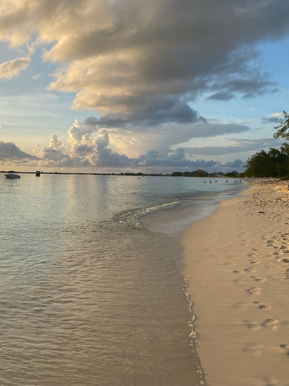 Calm beach at sunset with gentle waves, a sandy shoreline, footprints, boats on the horizon, and dramatic clouds in a serene sky.