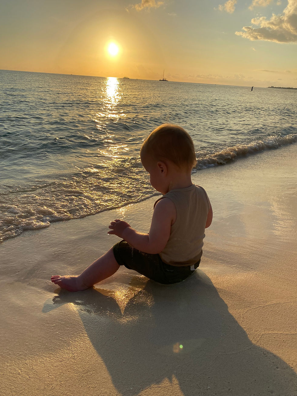 Toddler sitting on a sandy beach during sunset, watching gentle waves. A calm sea and sun with a sailboat silhouette in the background.