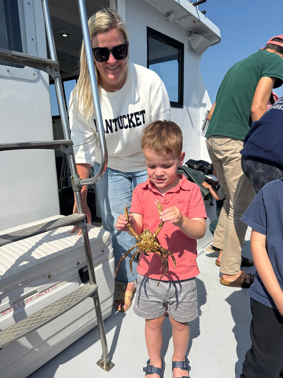 A woman in sunglasses smiles as a young boy in a red shirt holds up a crab on a boat. The word "NANTUCKET" is on her sweater.