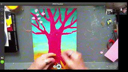 An artist uses pink pieces of paper to place atop of a vibrantly colored pink tree with a blue and orange background, done in acrylic paint.