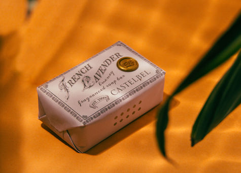 Product photo of a bar of soap on an orange background. The light looks like reflections from water and a few fronds from a plant lean into the frame 