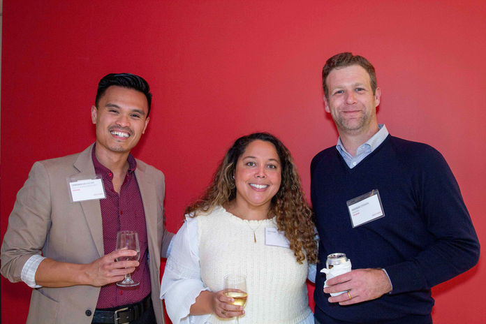 Portrait from an event. Three individuals stand together against a red wall, smiling at the camera