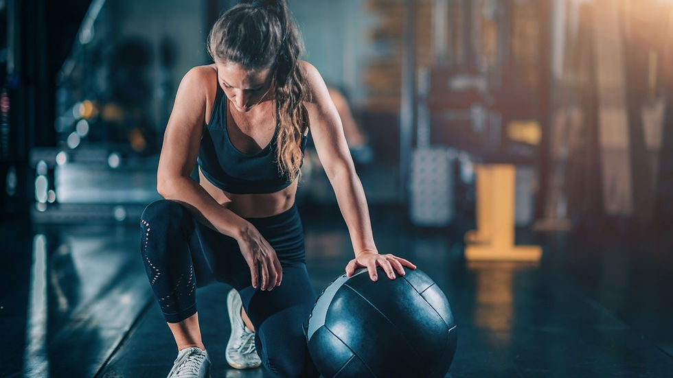 Woman kneels in a gym, resting a hand on a large medicine ball, looking down with a focused, tired expression.