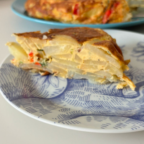 A colorful frittata filled with red peppers, salami, parsley, potatoes, onions, and eggs, served on a rustic plate. The dish is golden and fluffy, garnished with fresh herbs.