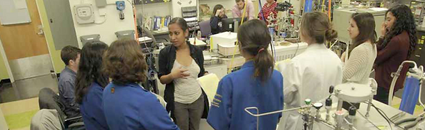 Aradhna Tripati is seen standing, in a laboratory. She is speaking with a group of 7 students who have formed a semi-circle in front of her. They are listening to Aradhna, facing away from the camera. Some are wearing labcoats. Lab machinery is seen in the foreground and background.