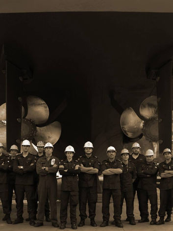 RNZN Naval Engineers standing in uniform in a dry dock below an RNZN Offshore patrol vessel with distinct propellers and service umbillicals seen running to the ship during a cruicial survey maintenance period