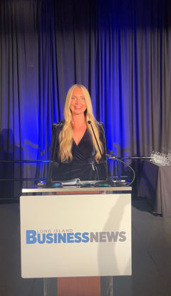 Courtney Chicvak at the Long Island Business News event, standing at a podium with the organization's logo. She is wearing a navy velvet dress with long blonde hair cascading over her shoulders. The background features blue-lit curtains and a table displaying glass awards.
