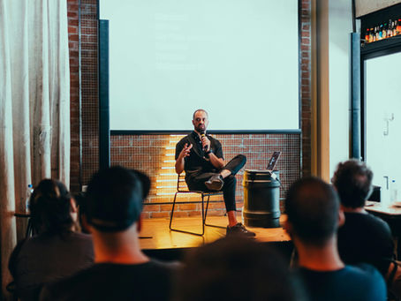 A speaker sits on a small stage under a large projector screen, speaking to an audience seated in a darkened seminar room during a professional presentation.