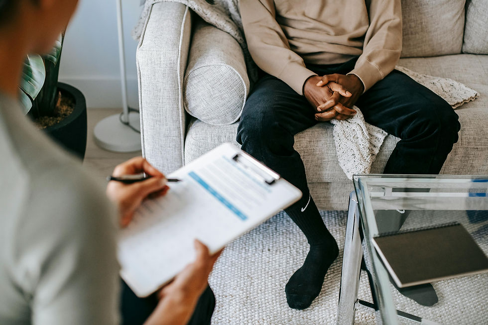 A person sitting on a couch with their hands folded with a person sitting in front of them with a clipboard filling out a questionnaire