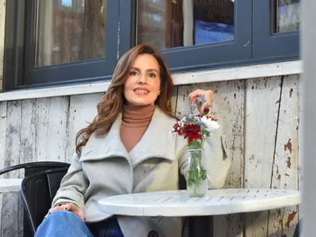 Omayra Jiménez posando sonriente en una mesa exterior con flores, luciendo un abrigo elegante y un estilo natural y cuidado.