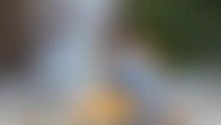 Omayra Jiménez sentada frente a una fuente de agua escalonada, concentrada en un cuaderno que sostiene. Viste una gabardina clara y jeans, y lleva unos llamativos zapatos con estampado de leopardo. El fondo es un muro de piedra y vegetación verde.