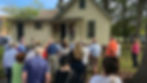 crowd behind a victorian house listening to a speaker in a black dress on the back porch.