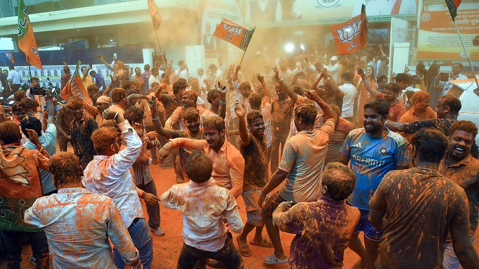 People covered in orange powder dance and celebrate, waving flags in a lively crowd. Banners are displayed in the background.