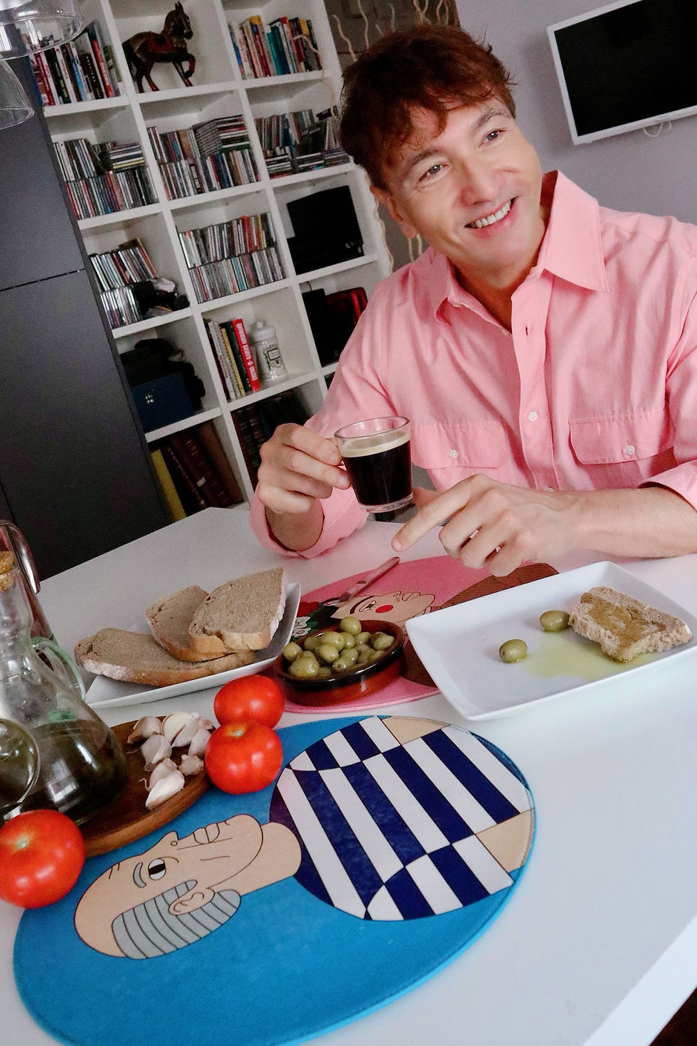 The Trendy Man sonriente con camisa rosa sostiene café en una mesa con pan, aceitunas, tomates y ajo; salón con estantería.