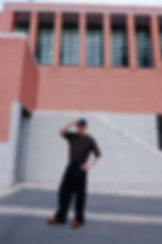 The Trendy Man saluda con gorra azul frente a edificio de ladrillo rojo. Viste camiseta oscura de Uniqlo y pantalones negros también de Uniqlo. Fondo urbano moderno.