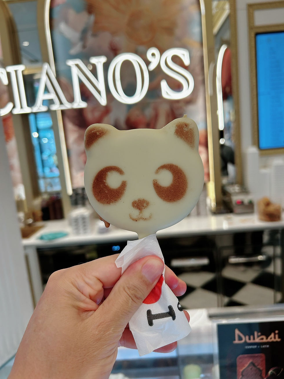 Mano sostiene una paleta de panda blanco y café en una heladería; al fondo se lee LUCIIANO'S y el ambiente es brillante.