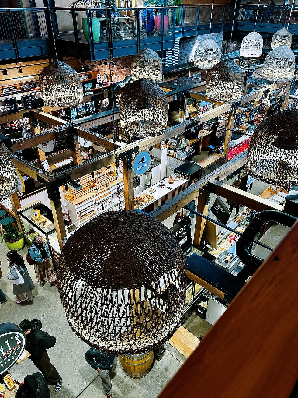 Mercado interior de Christchurch, New Zealand, con lámparas colgantes de mimbre; personas compran y caminan entre puestos, carteles Bellbird y BBQ.