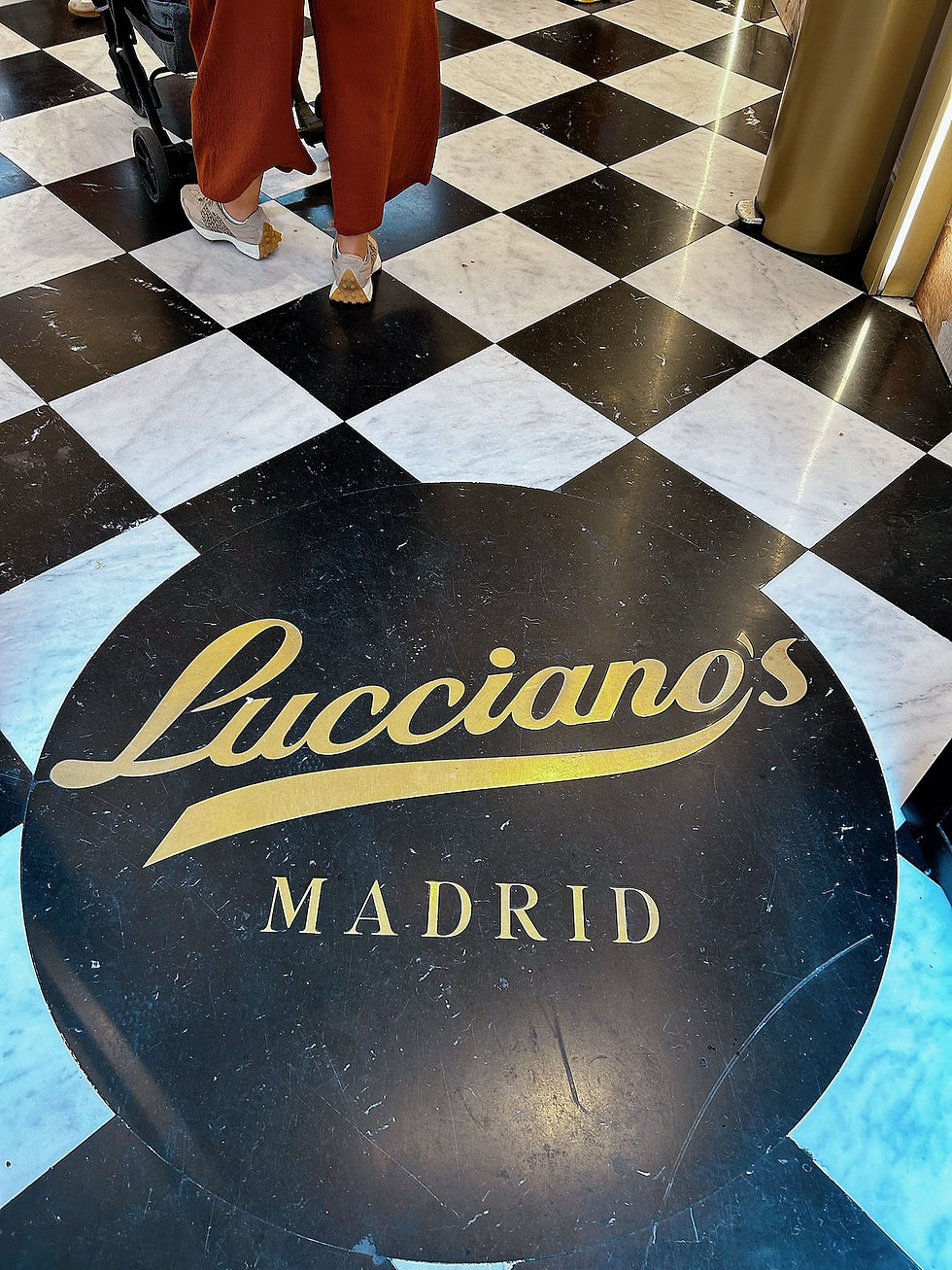 Persona con pantalón rojo y zapatillas junto a un carrito sobre suelo ajedrezado; logo Lucciano's MADRID en un círculo negro.