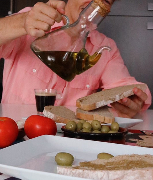 The Trendy Man en su casa con camisa rosa vertiendo aceite de oliva sobre pan en una mesa con tomates, aceitunas y un vaso.