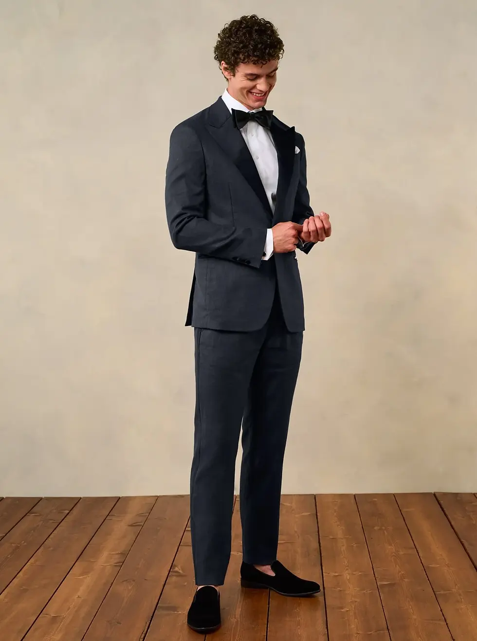 Novio con traje oscuro y pajarita, ajusta puño mientras sonríe. Fondo neutro y suelo de madera, ambiente elegante.