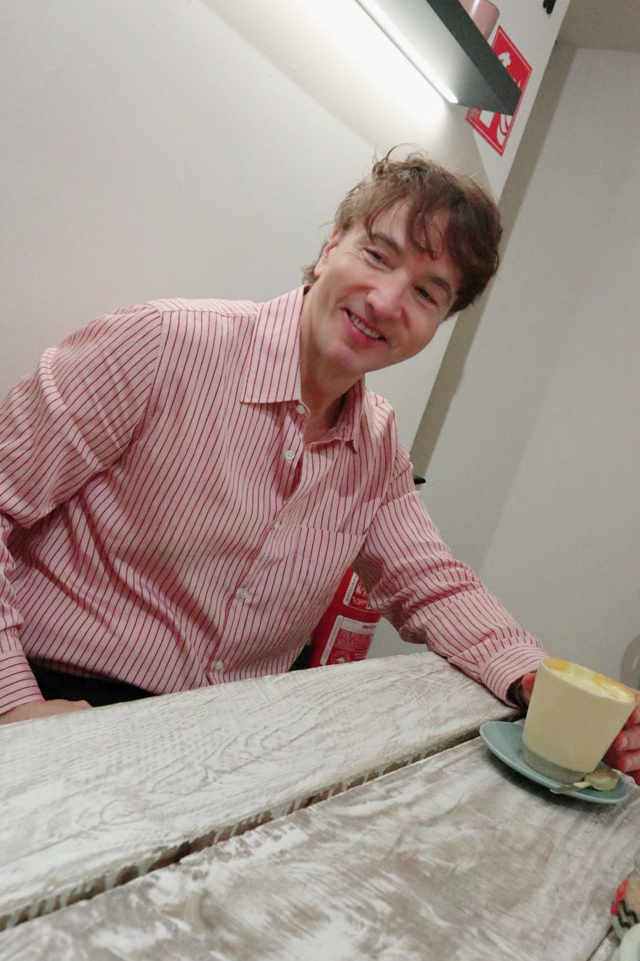 The Trendy Man  sonríe en una mesa de curcuma latte, con camisa a rayas rojas. Sostiene una taza de café con leche. Fondo de pared clara y extintor rojo.