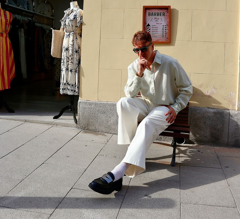 The Trendy Man con gafas de sol y ropa clara sentado en banco frente a tienda con maniquí. Cartel de "Barber Shop" en la pared. Ambiente soleado en la calle Noviciado de Madrid