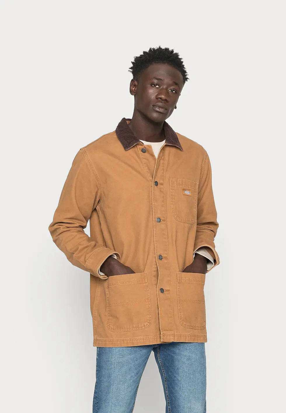 Hombre con chaqueta beige y vaqueros azules, de pie con manos en los bolsillos sobre fondo blanco; expresión serena.