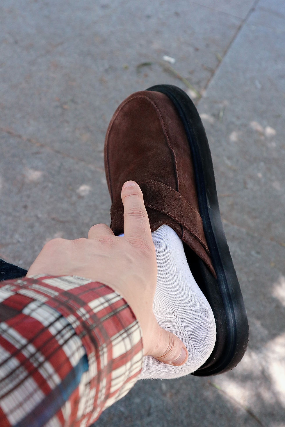 The Trendy Man con camisa a cuadros ajusta un zapato marrón sobre calcetín blanco en el pie, fondo de suelo gris.