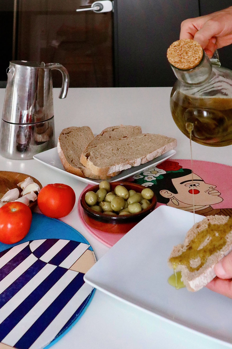 Manos sirven aceite de oliva sobre pan en mesa con tomates, aceitunas, ajo y cafetera, en ambiente casero.